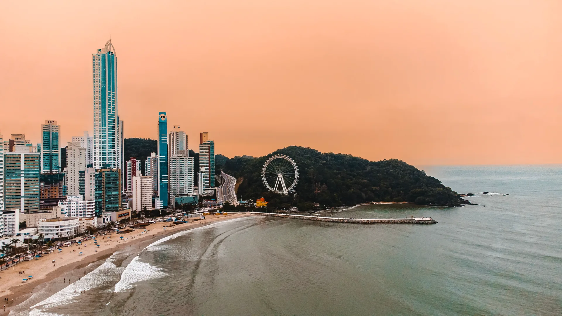Vista aérea do centro de Balneário Camboriú ao entardecer, com prédios altos à beira-mar e a roda-gigante do Unipraias ao fundo.